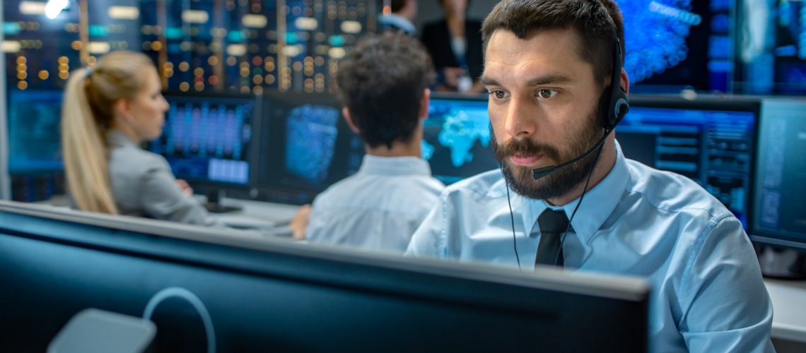 Customer Service Representative Answer Client's Questions in a Headset. He Works for a Big Technological Company. Office Has Multi-Ethnic Team of Specialists, Displays Show Useful Information.